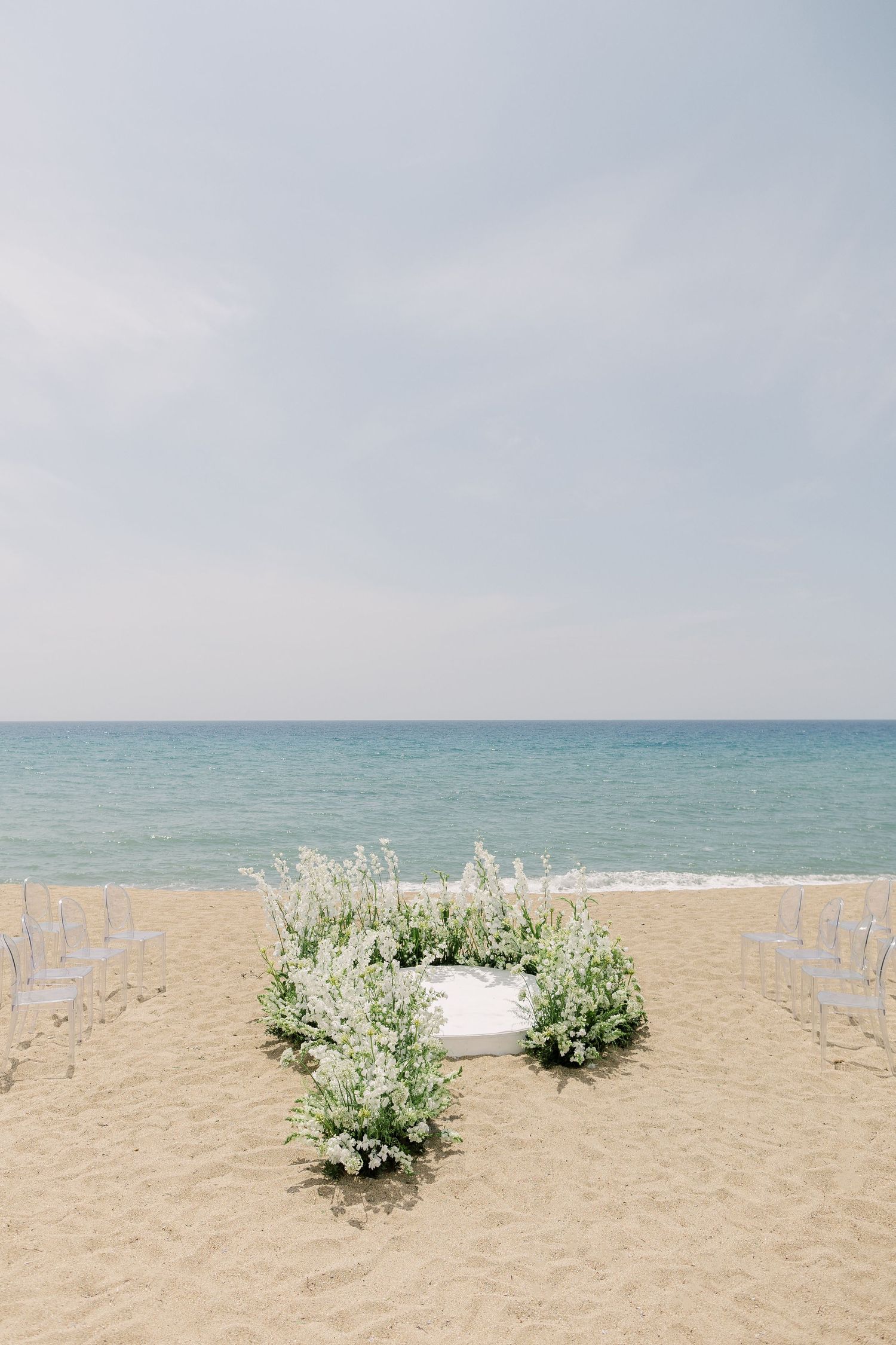 White wooden chairs arranged on a sandy beach with green floral arrangements and ocean views create a romantic ceremony setting.