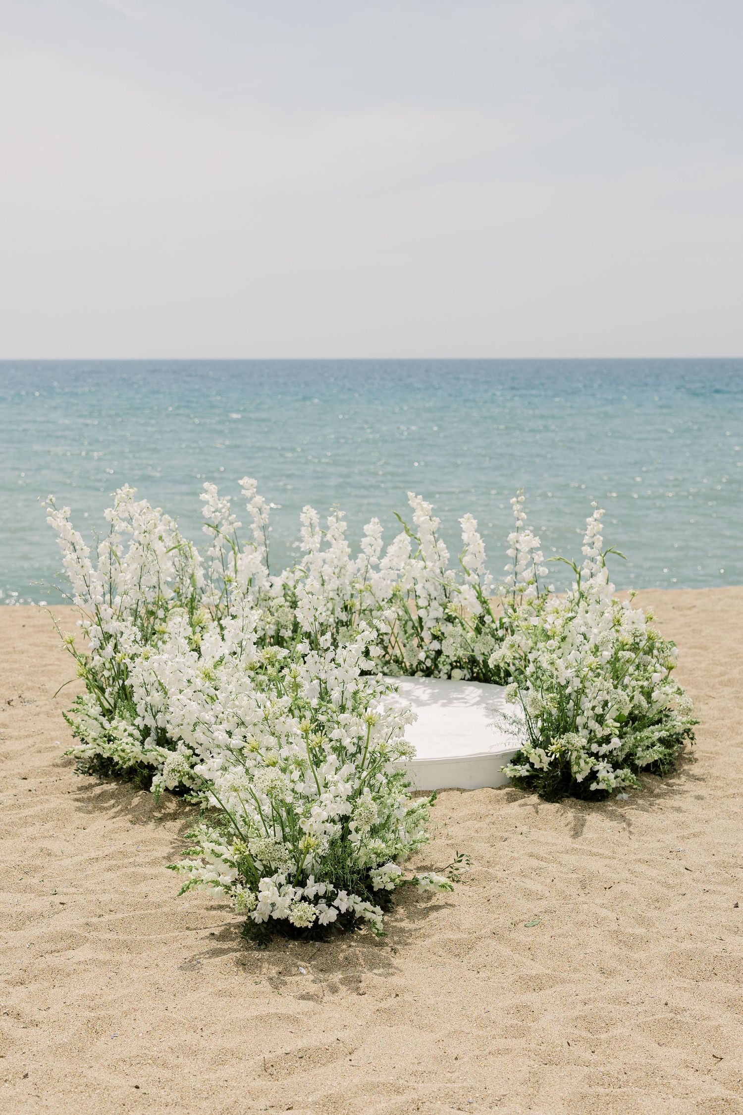 White floral arrangements surround a cushion on a sandy beach with turquoise ocean waters in the background.