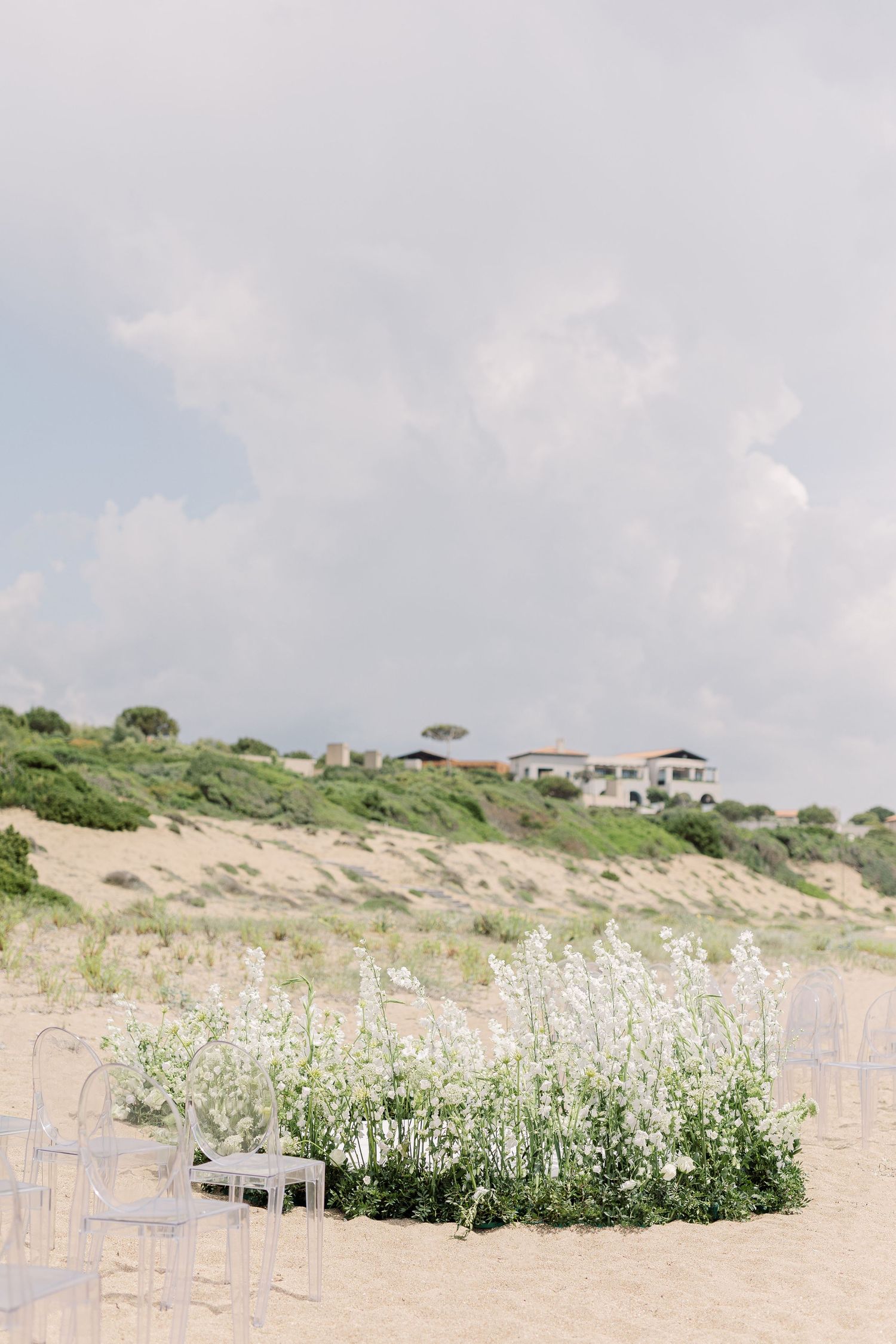 White wooden chairs arranged in rows on a sandy beach with coastal vegetation and cloudy skies in the background.