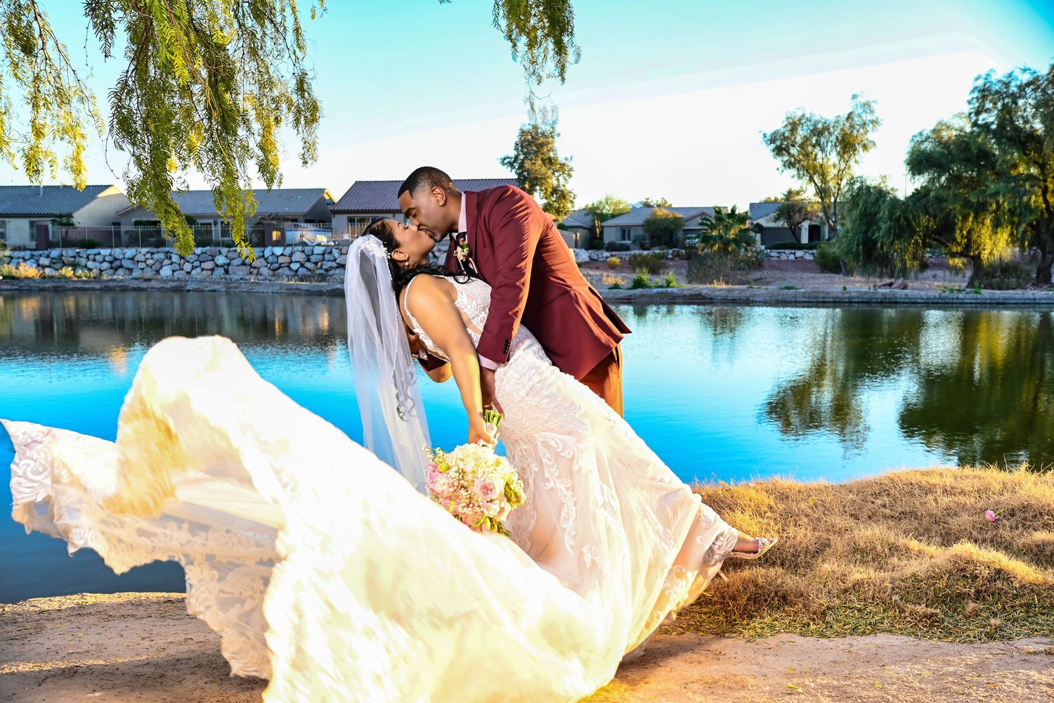 Bride and groom sharing a romantic kiss by a serene lake, with her flowing white gown billowing in the breeze.