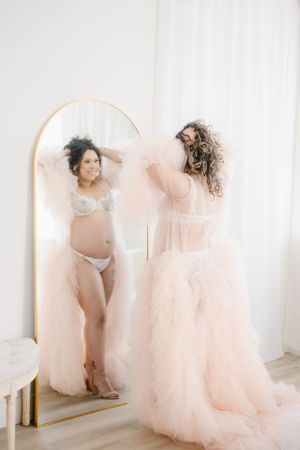Artistic maternity mirror pose with flowing pink tulle dress against white backdrop.