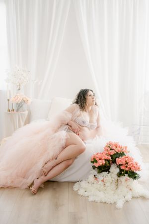Elegant maternity pose on white couch with pink flowers and flowing tulle gown.