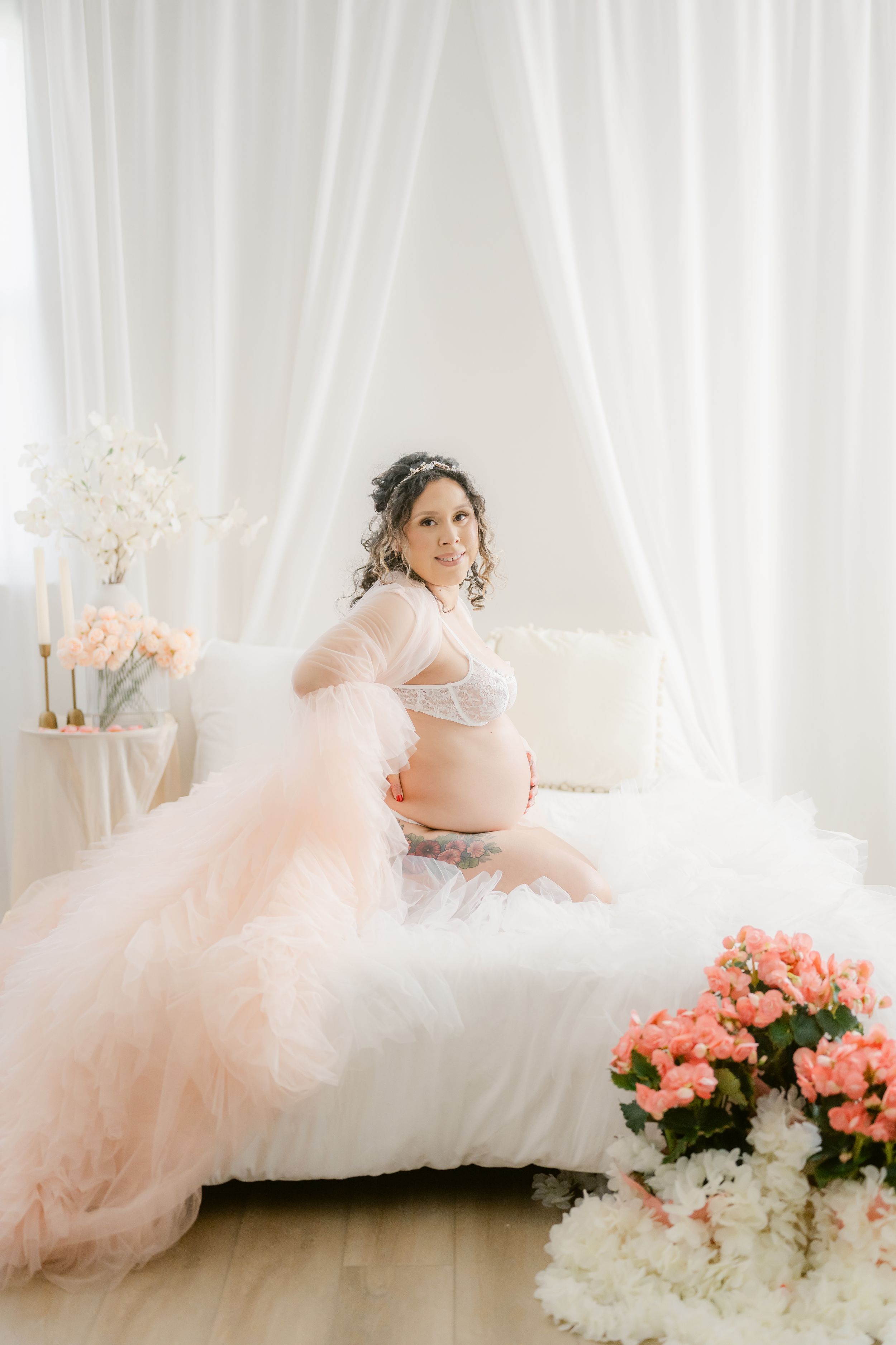 Dreamy maternity photo with pink tulle gown and floral arrangements in bright white room.