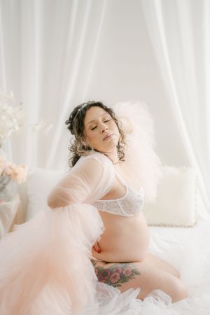 Ethereal maternity boudoir photo with flowing pink tulle and white lace lingerie.