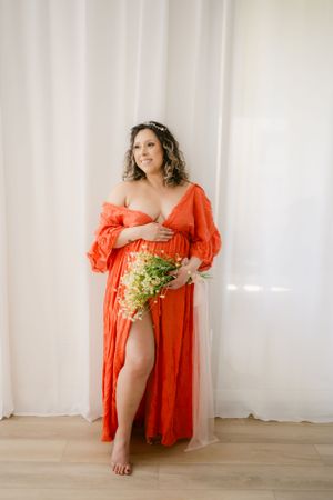 Elegant maternity portrait in flowing coral dress holding fresh flowers against white backdrop.