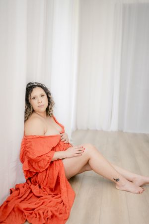 Artistic maternity portrait in coral dress sitting against white curtains.