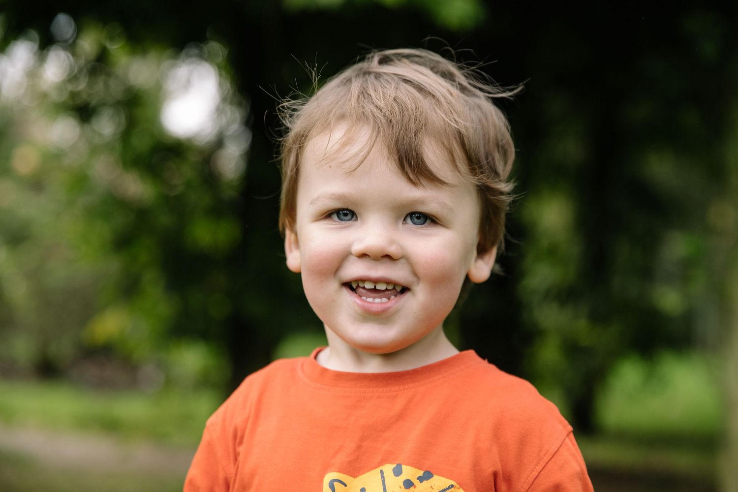 A child wearing an orange t-shirt smiles brightly outdoors against a green background.