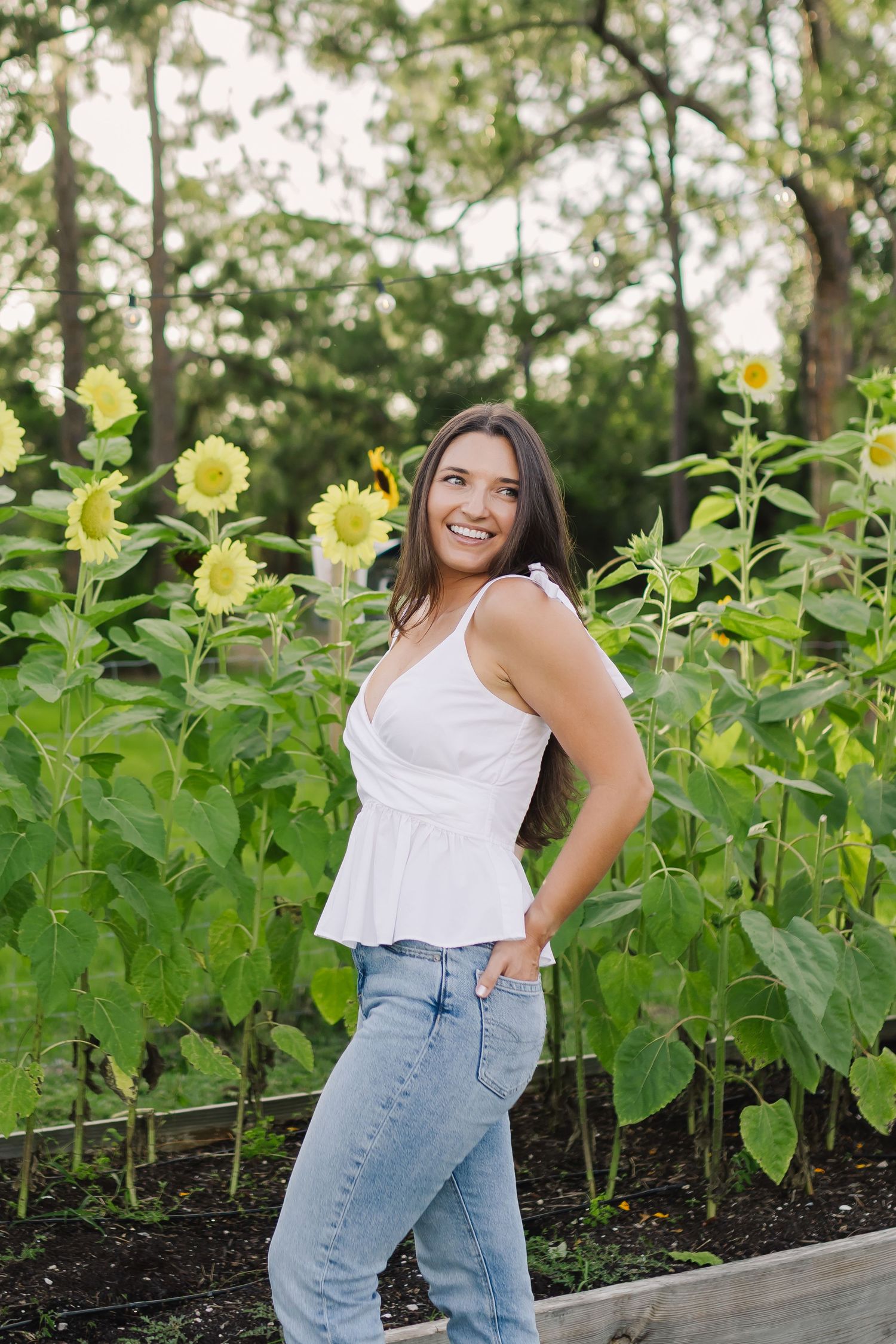 Smiling young woman in white top and jeans poses among tall sunflowers in a garden.