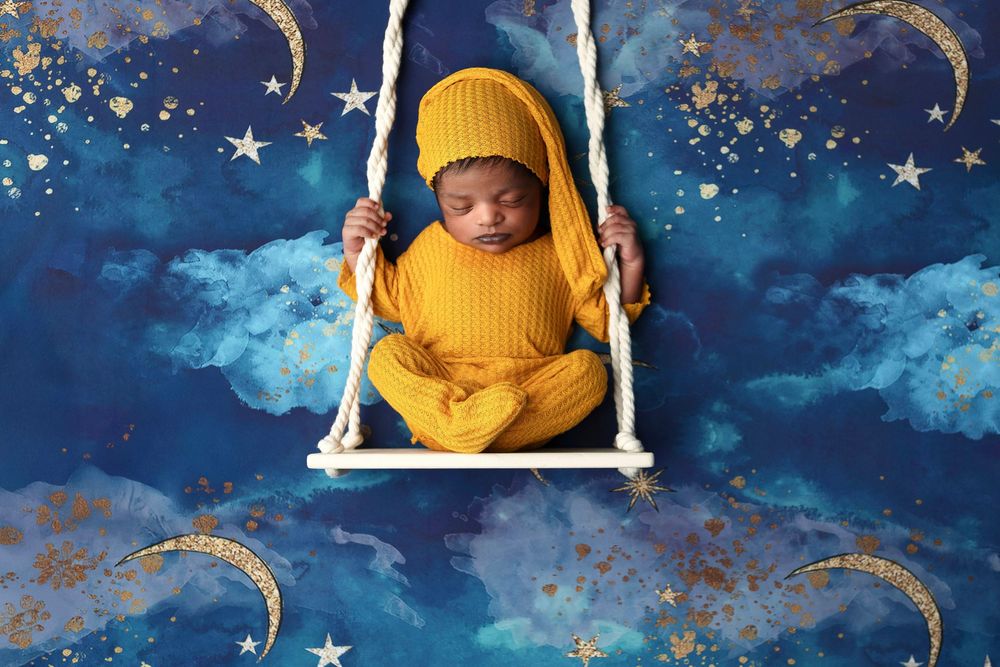 Newborn baby in yellow outfit sleeping on a white swing against a dreamy blue starry night sky backdrop.