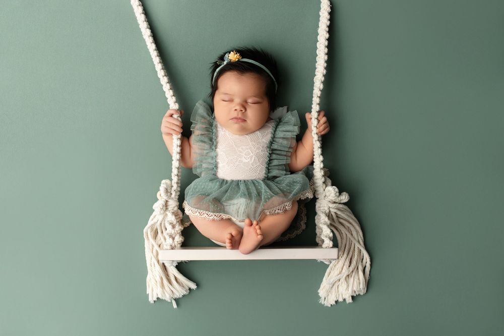 Sleeping baby in tulle skirt resting on white rope swing against sage green wall.