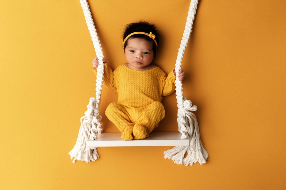 Baby in yellow knit outfit sitting on white rope swing against bright yellow background.