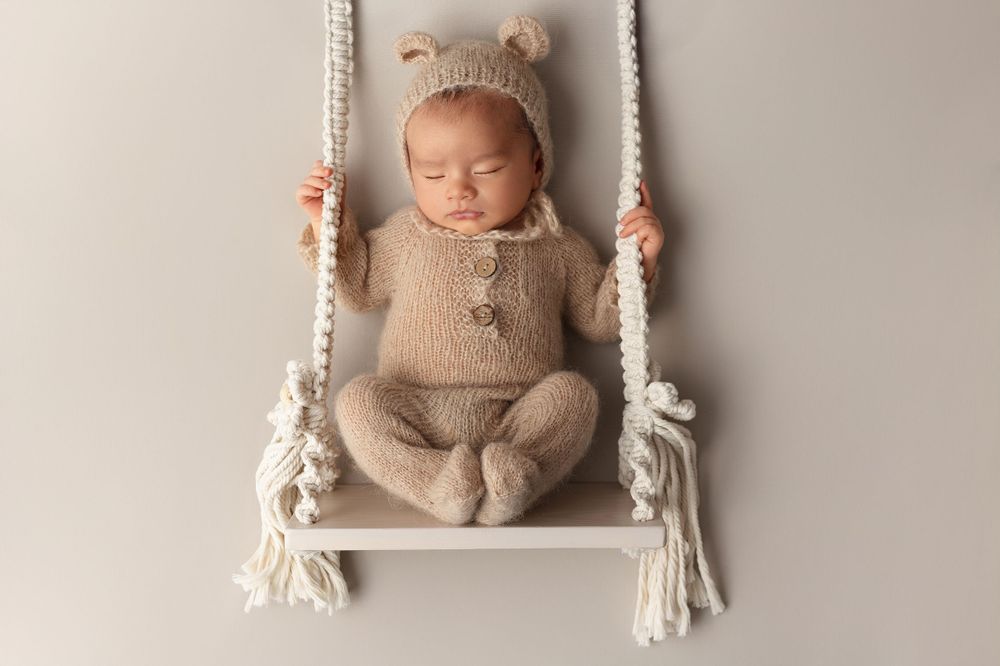 Sleeping newborn in beige knit outfit resting on white macrame swing.