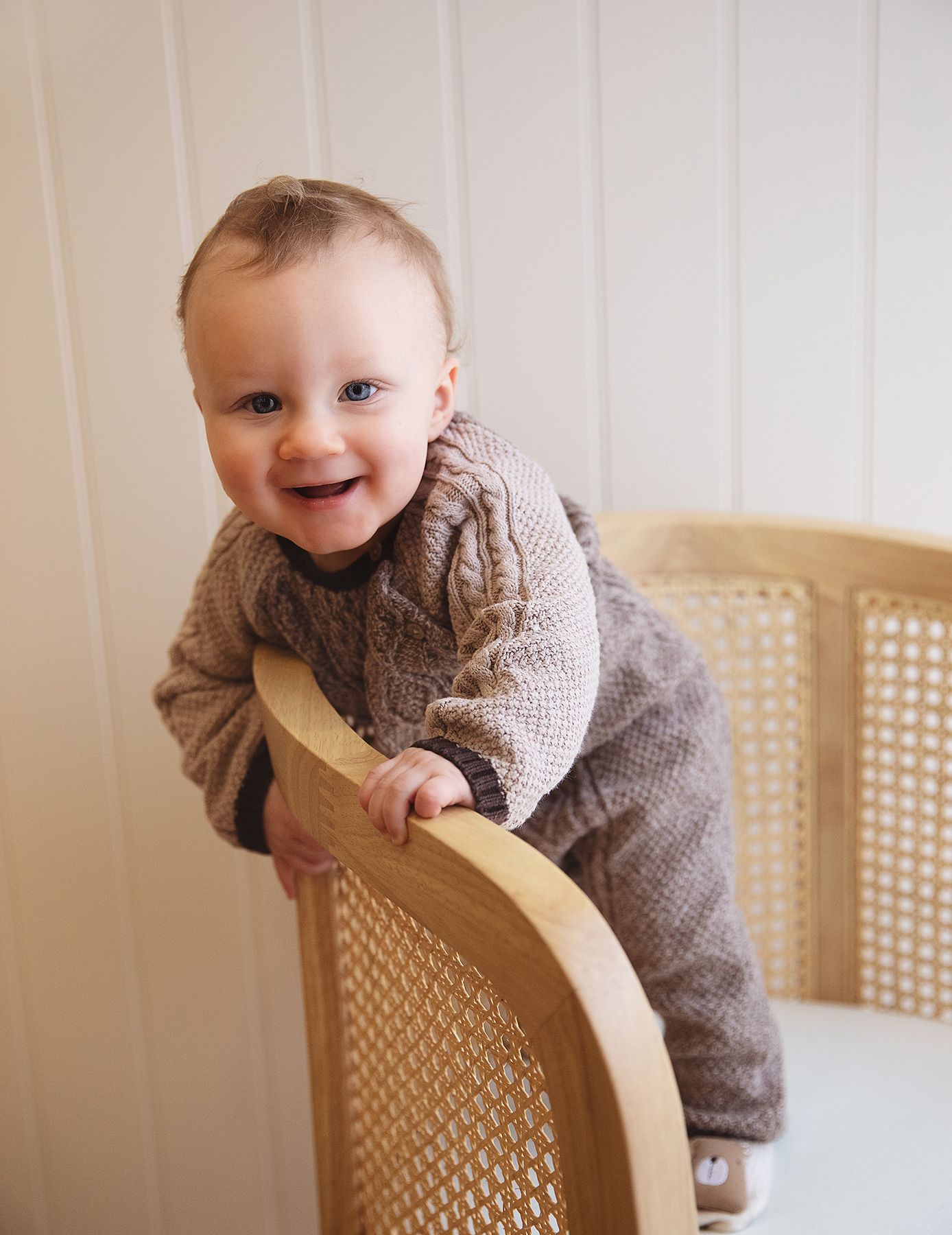 Baby wearing grey knit sweater smiles while leaning on cane-backed dining chair in bright interior setting. During a photoshoot in Bexley