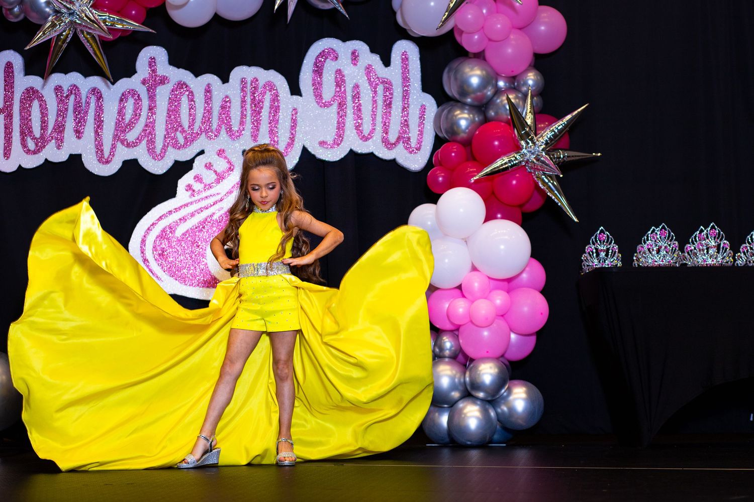 Performer in flowing yellow gown twirls dramatically on stage with pink balloon arch and hometown decor.