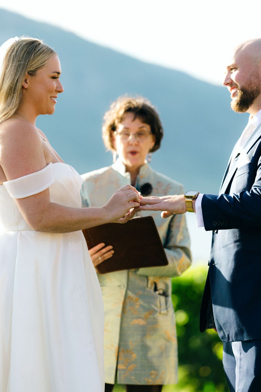 Wedding ceremony in progress against mountain backdrop as couple exchanges rings. Modern Wedding in the Okanagan