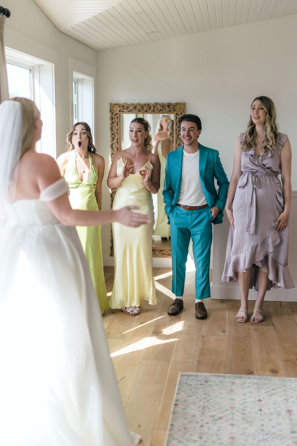 Wedding party members in colorful dresses and suits react with joy as they see the bride in her white gown.