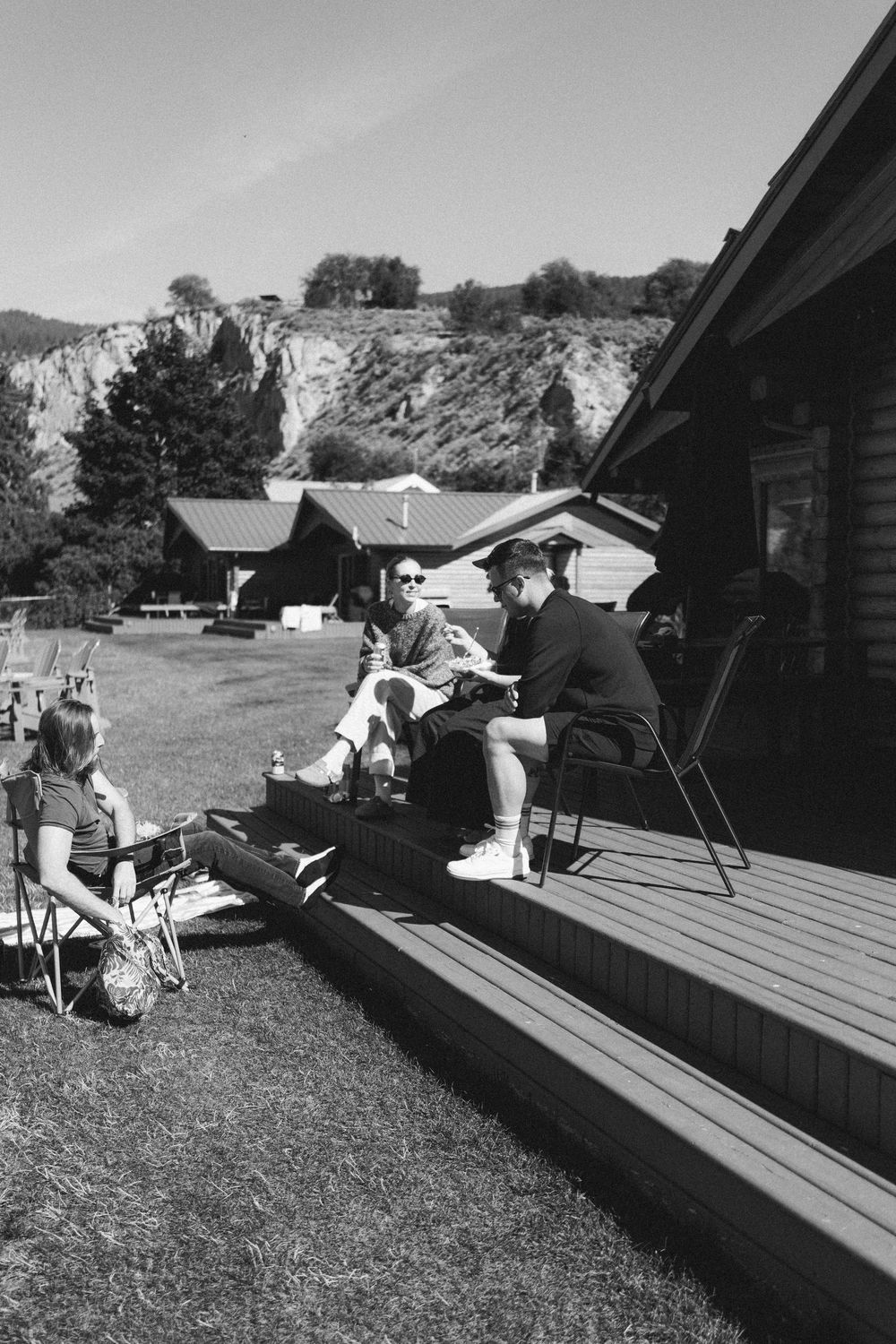 Black and white photo of people relaxing on a cabin deck in a scenic mountain setting.