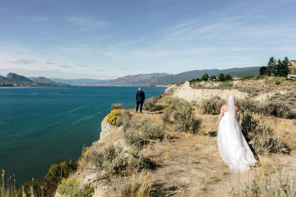 Bride and groom stand on cliffside overlooking dramatic blue lake and mountain landscape.