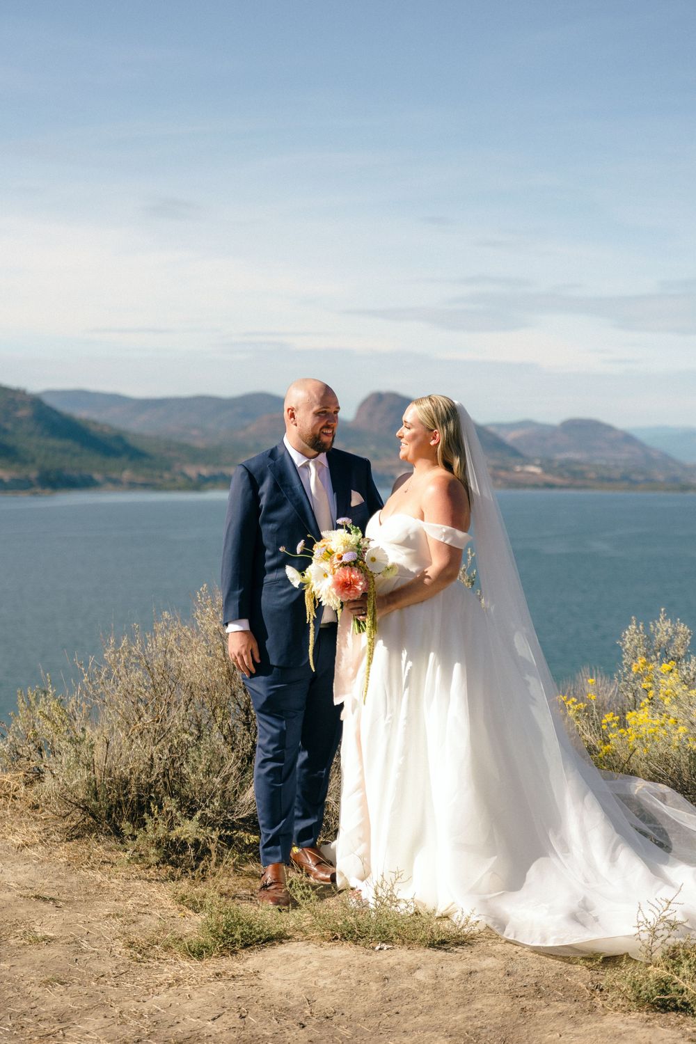 A couple poses for a portrait on a cliffside overlooking a lake and mountain landscape.Poplar Grove winery Modern Wedding in the Okanagan