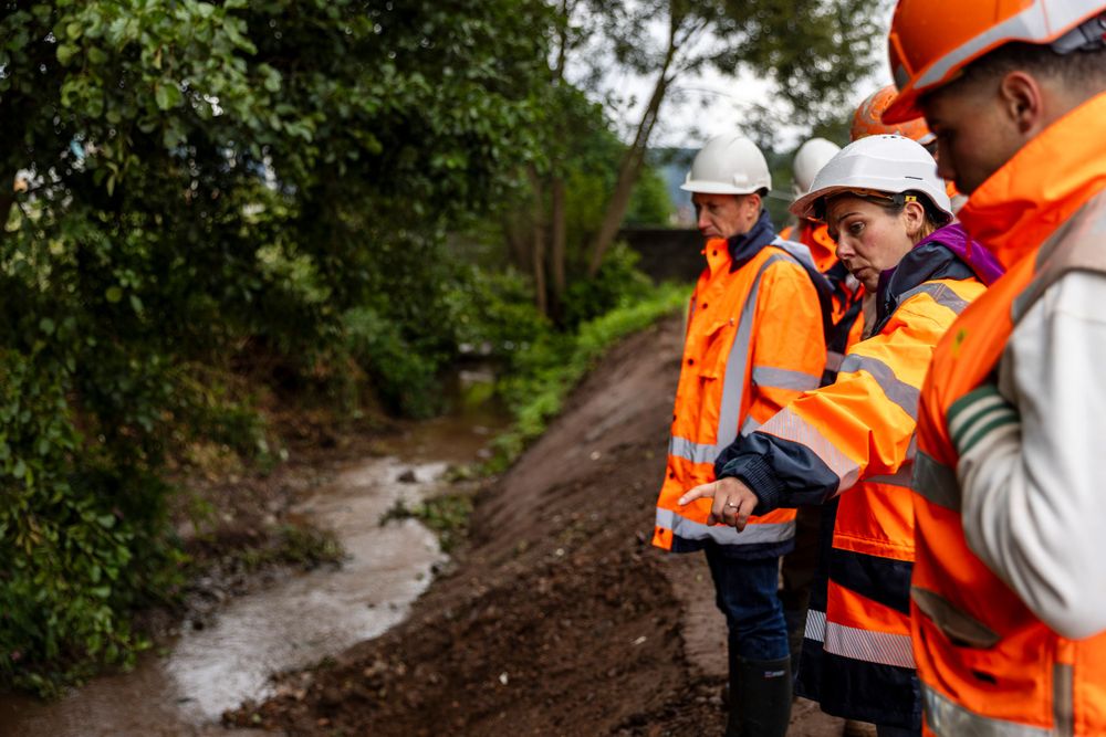 Construction workers in orange safety vests inspect a muddy stream bank at an outdoor worksite.
