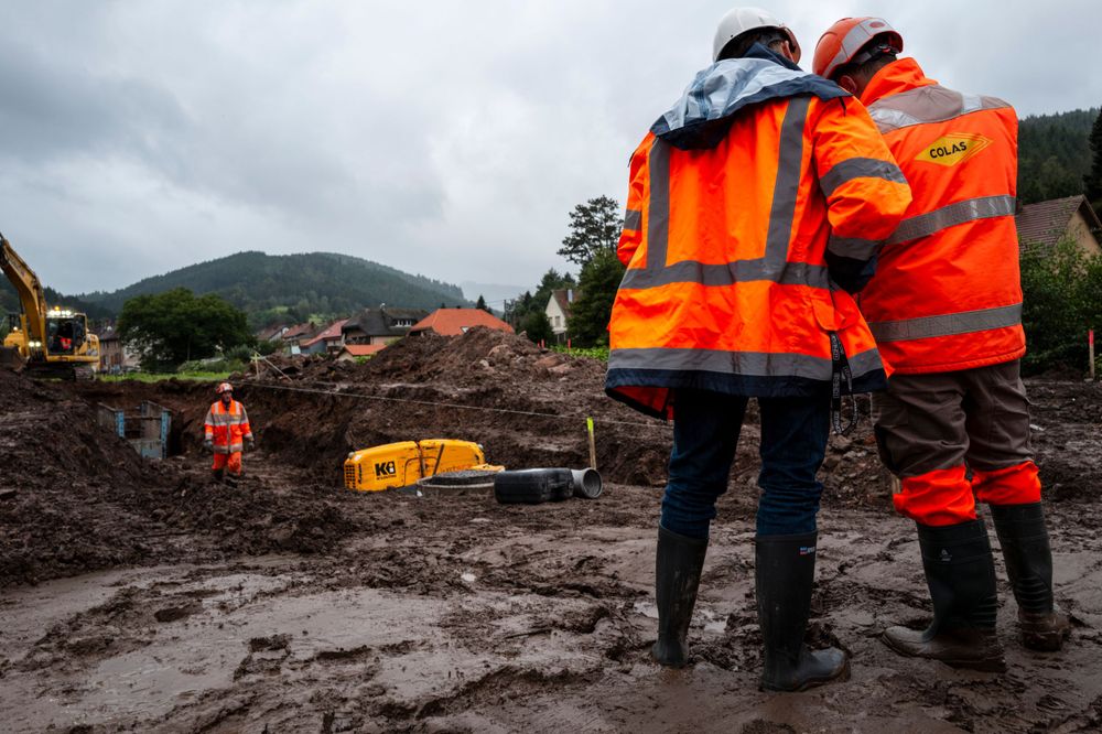 Workers in orange safety vests observe muddy construction site in mountainous area.
