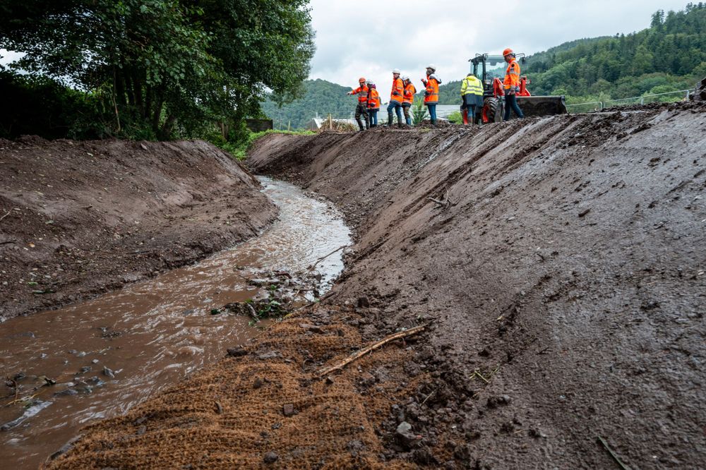 Group of workers in safety gear inspect muddy drainage channel on hillside.