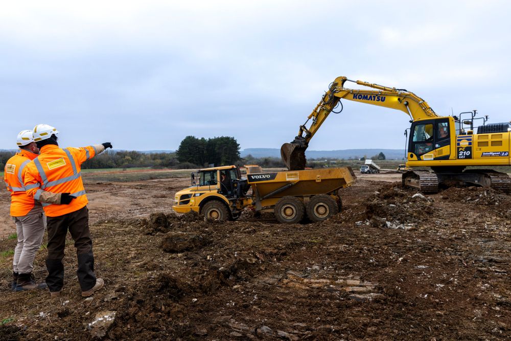 Construction workers in orange safety vests supervise excavator and dump truck operations at a dirt site.
