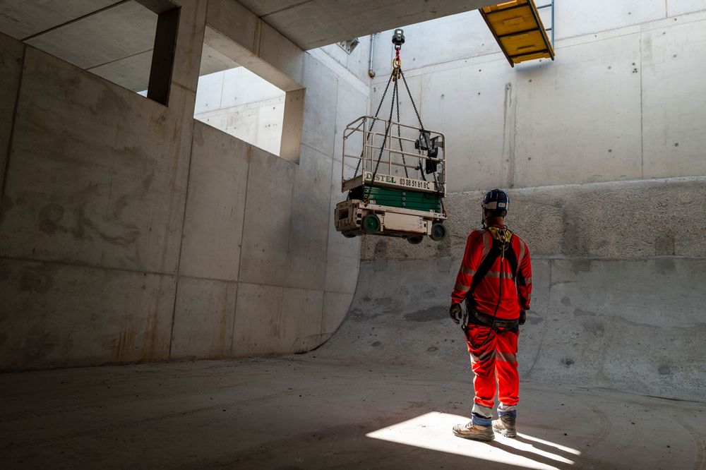 Construction worker in red safety gear stands in concrete tunnel as heavy equipment is lifted by crane overhead.