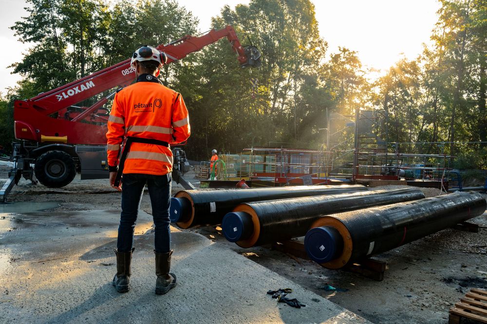 Construction worker in orange safety gear oversees large pipeline installation at an industrial site with a crane at sunrise.