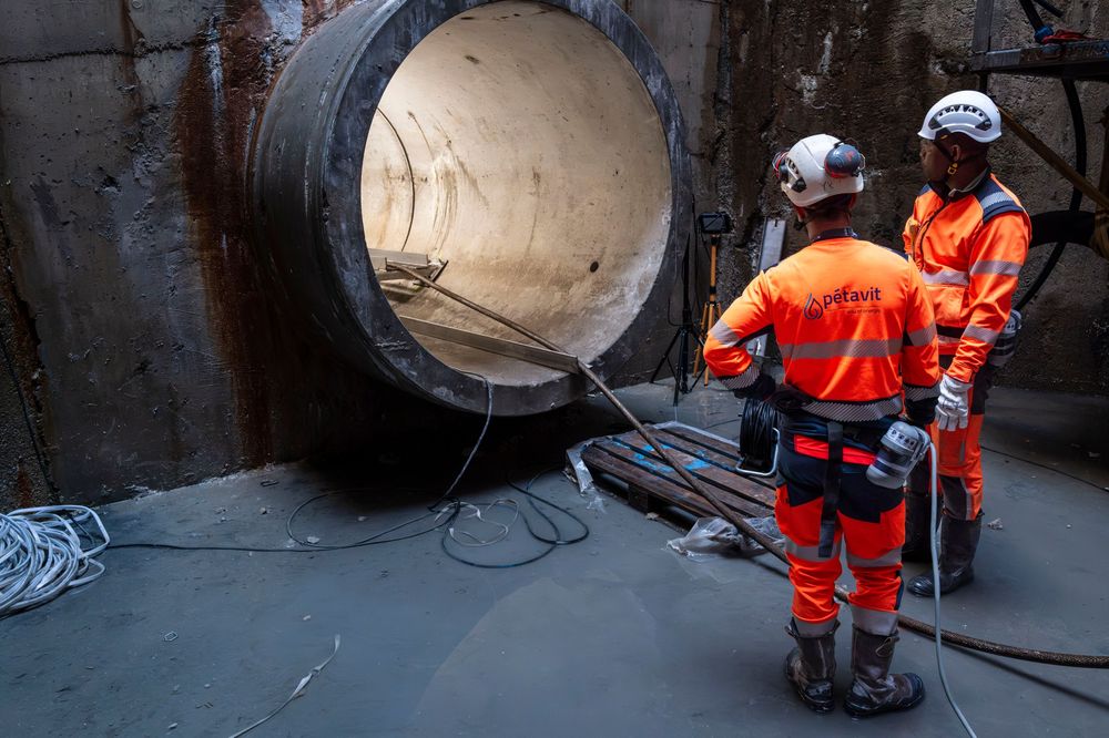Construction workers in orange safety gear inspect a large concrete tunnel entrance at an underground infrastructure site.
