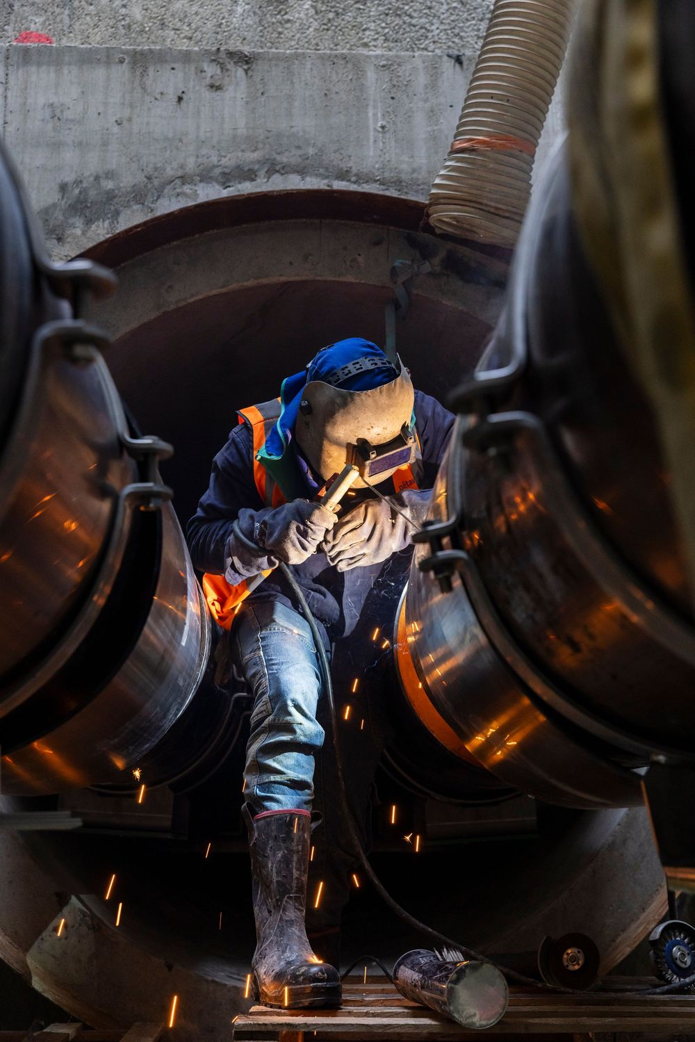 Welder in protective gear and helmet works between large metal pipes with sparks flying in industrial setting.