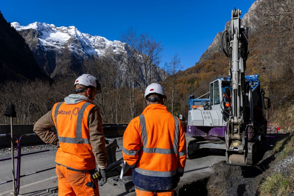 Workers in orange safety vests inspect a mountainous construction site with snow-capped peaks in the background.