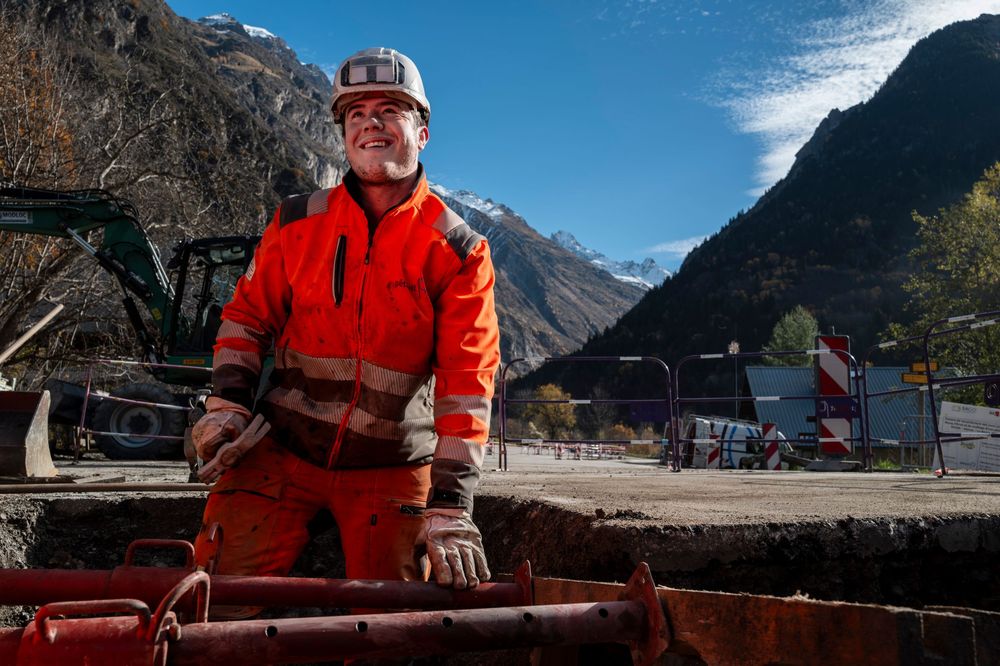 Scenic mountain landscape with snow-capped peaks and a person in bright orange safety gear standing near a railing.