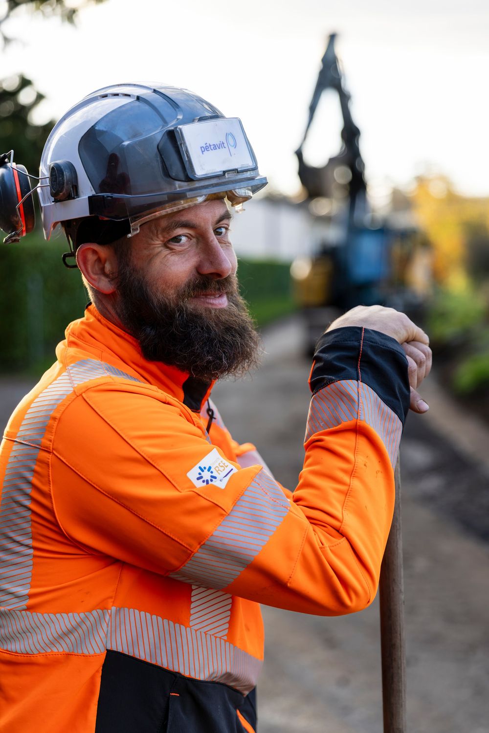 Construction worker in high visibility orange safety jacket hammers a stake into the ground at a worksite.