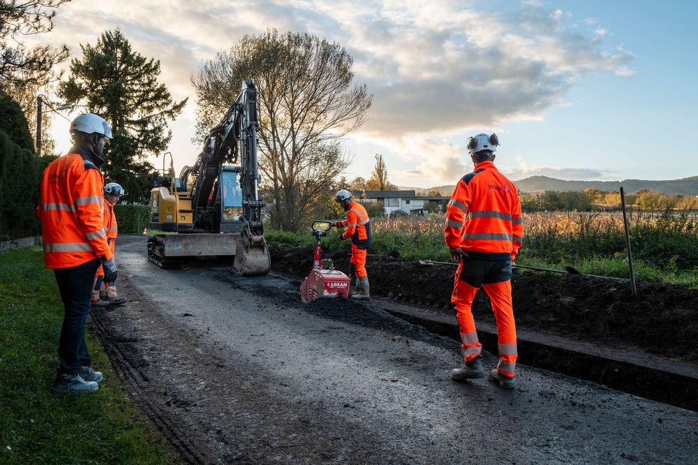Workers in high visibility orange safety gear walk along a rural pathway during a cloudy day.