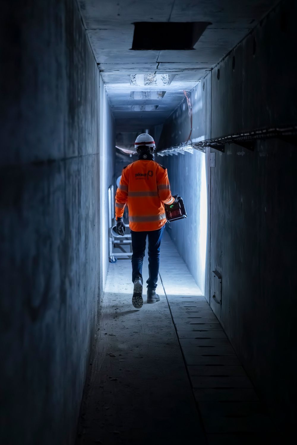 A person in an orange safety vest walks through a dimly lit concrete tunnel carrying inspection equipment.