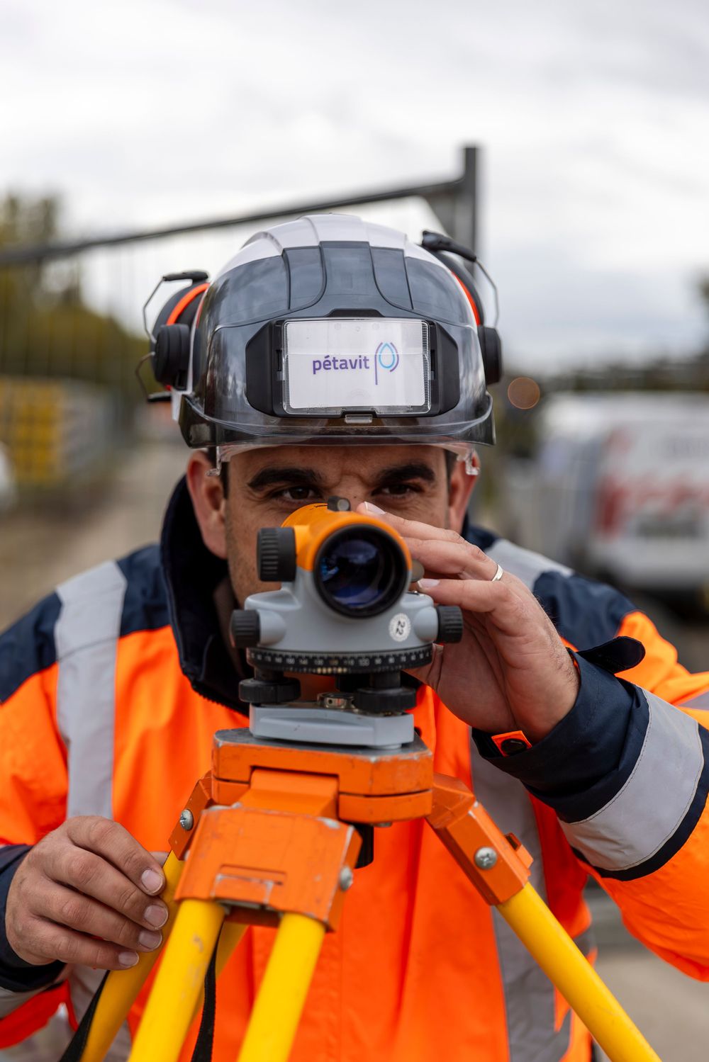 Surveyor in bright orange safety vest using professional surveying equipment with tripod and measuring device.
