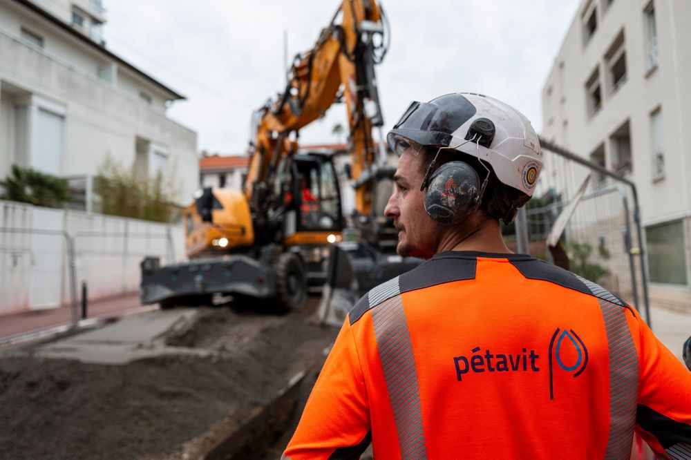 Construction worker in orange safety vest observes excavator at urban work site.