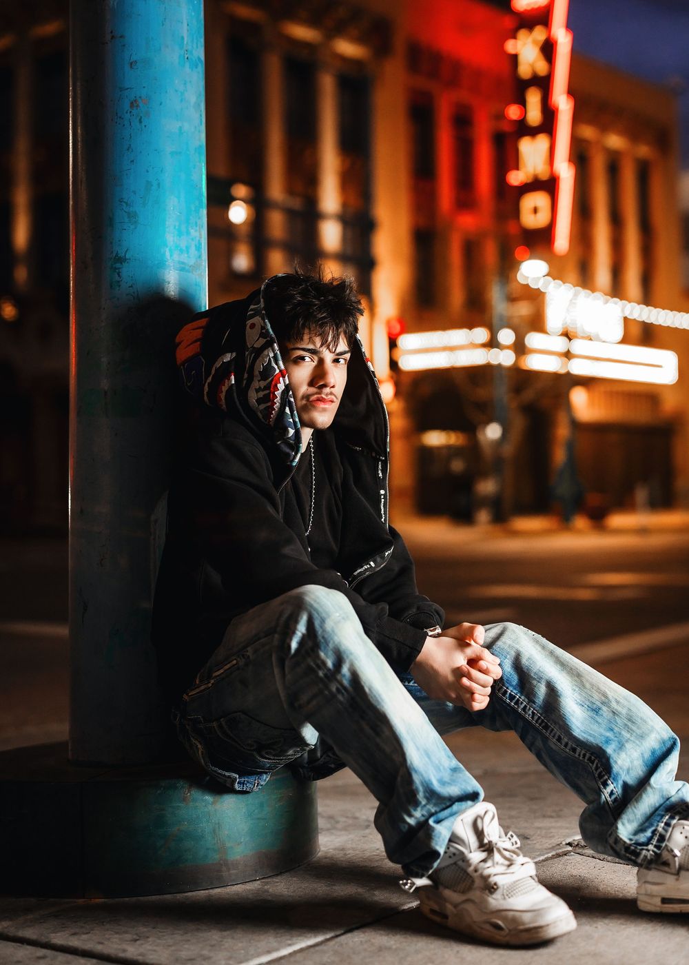 Young man in black hoodie and jeans sitting against a blue pole on a city street at night near a lit theater.