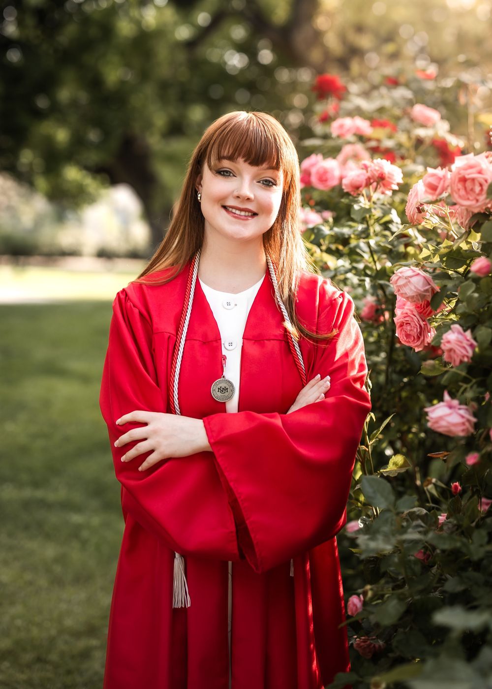 A graduate in bright red academic regalia stands smiling near blooming pink roses in a sunny garden setting.