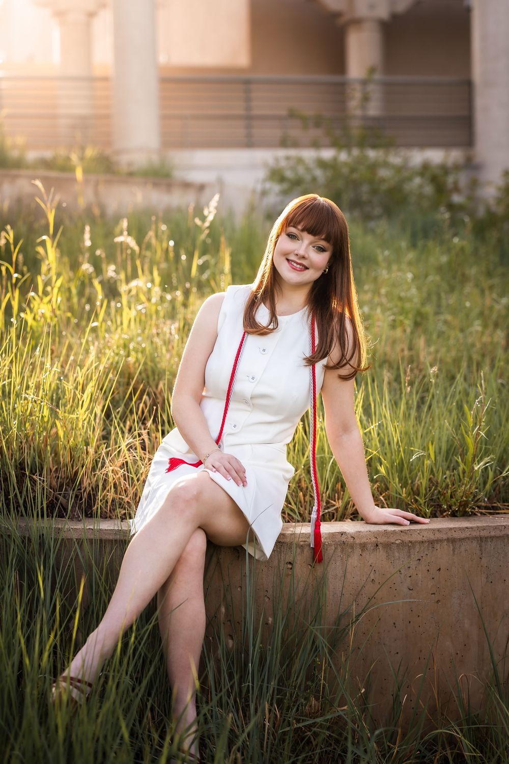 A figure in a white dress sits on a concrete ledge surrounded by tall grass during golden hour.