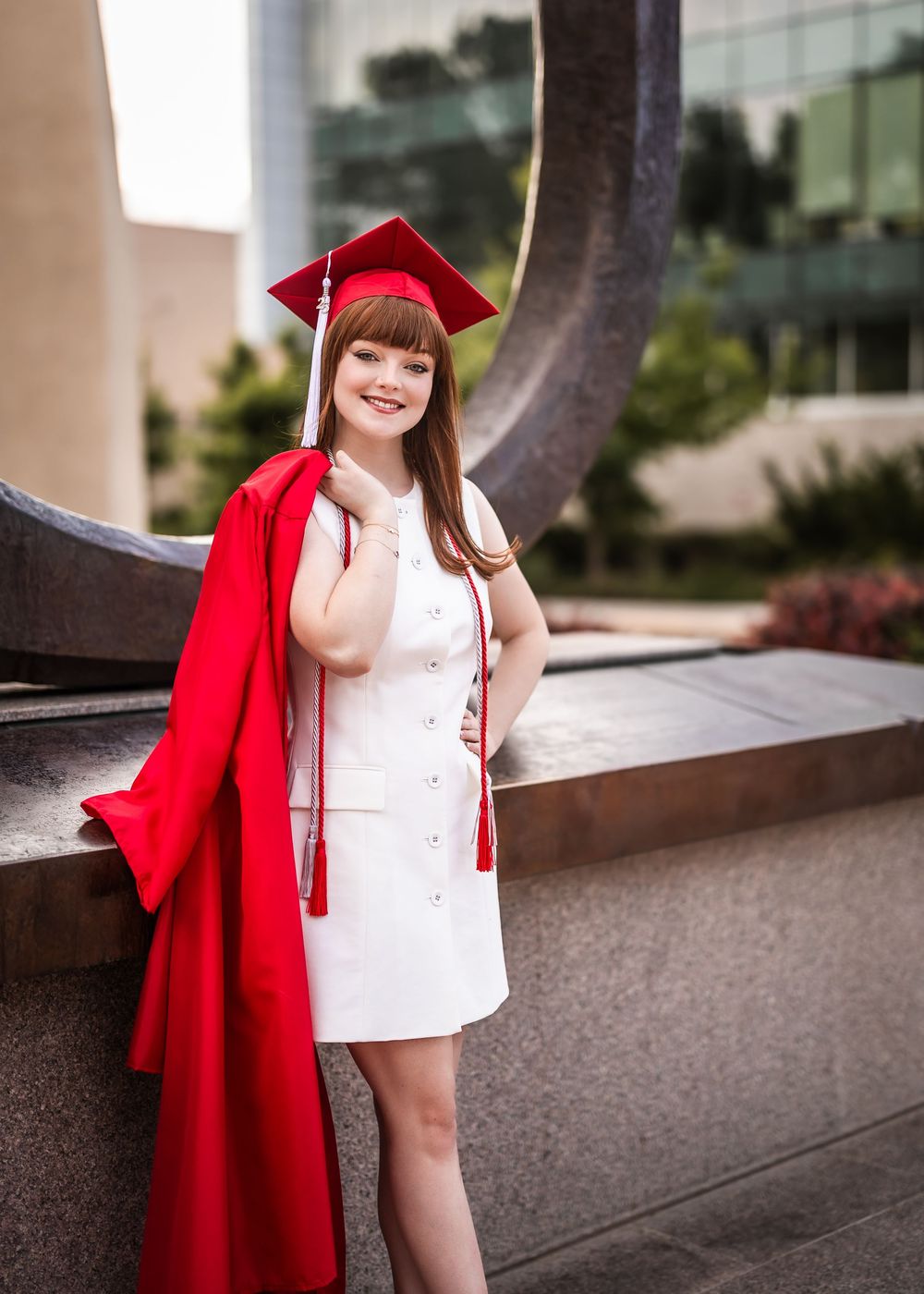 Graduate in a vibrant red cap and gown poses next to modern architecture during graduation photos.