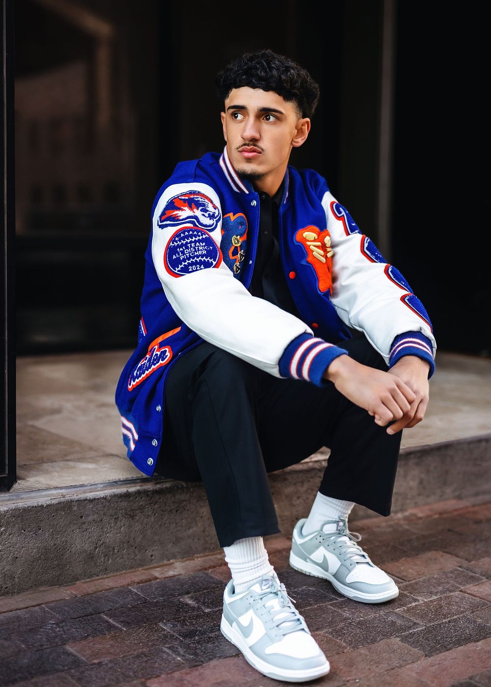 Young man in blue varsity jacket, black pants, and gray Nike Dunks sits on steps in urban setting.
