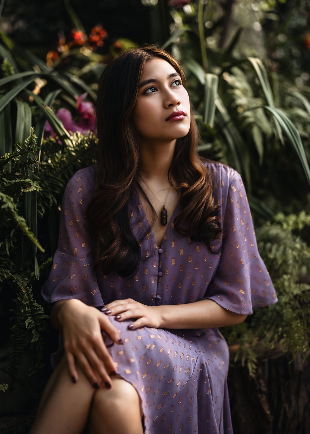 Young woman in purple floral dress sitting among lush tropical greenery, gazing upward thoughtfully.