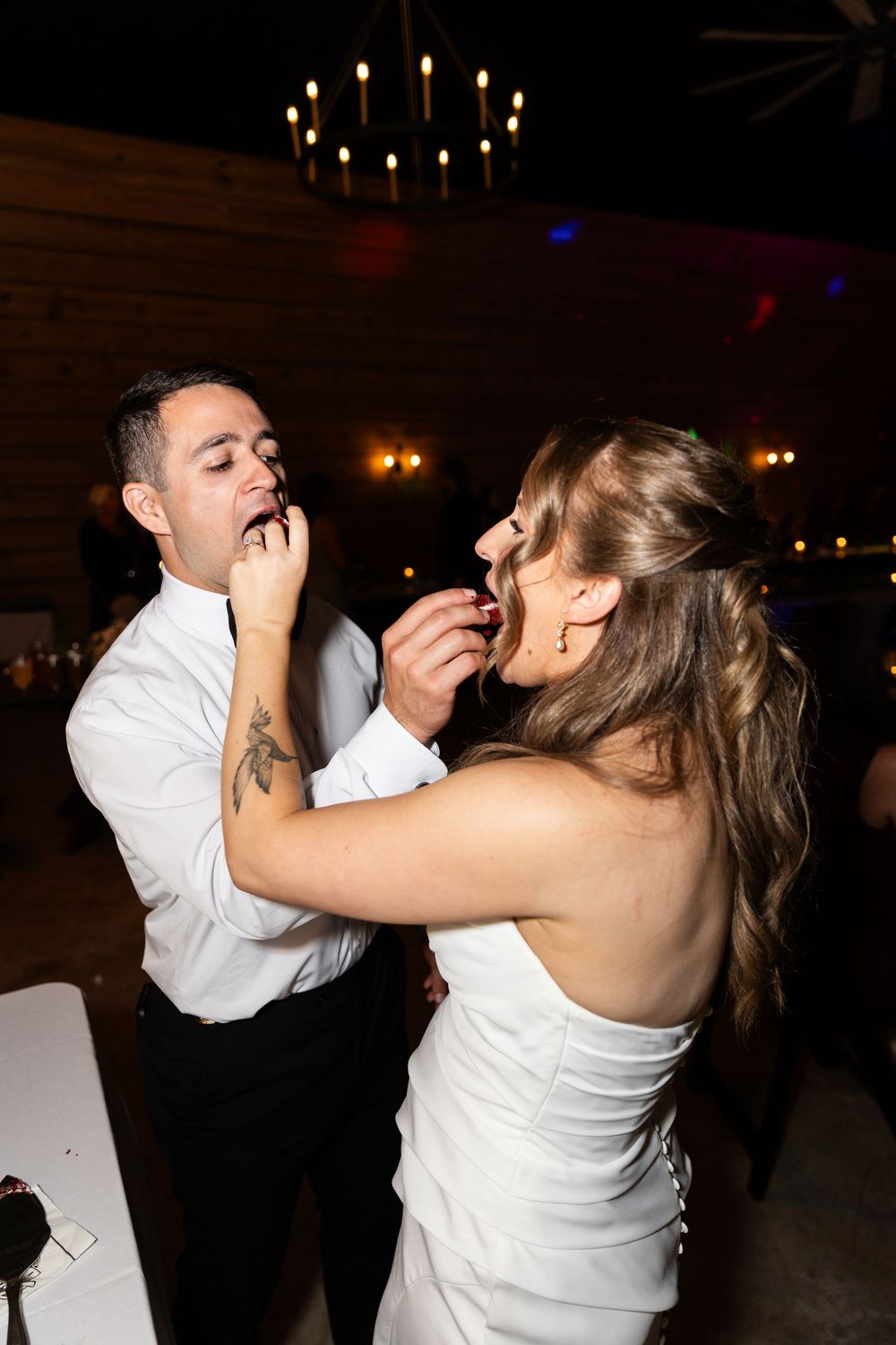 A bride and groom playfully feed each other cake at their wedding reception under a chandelier.