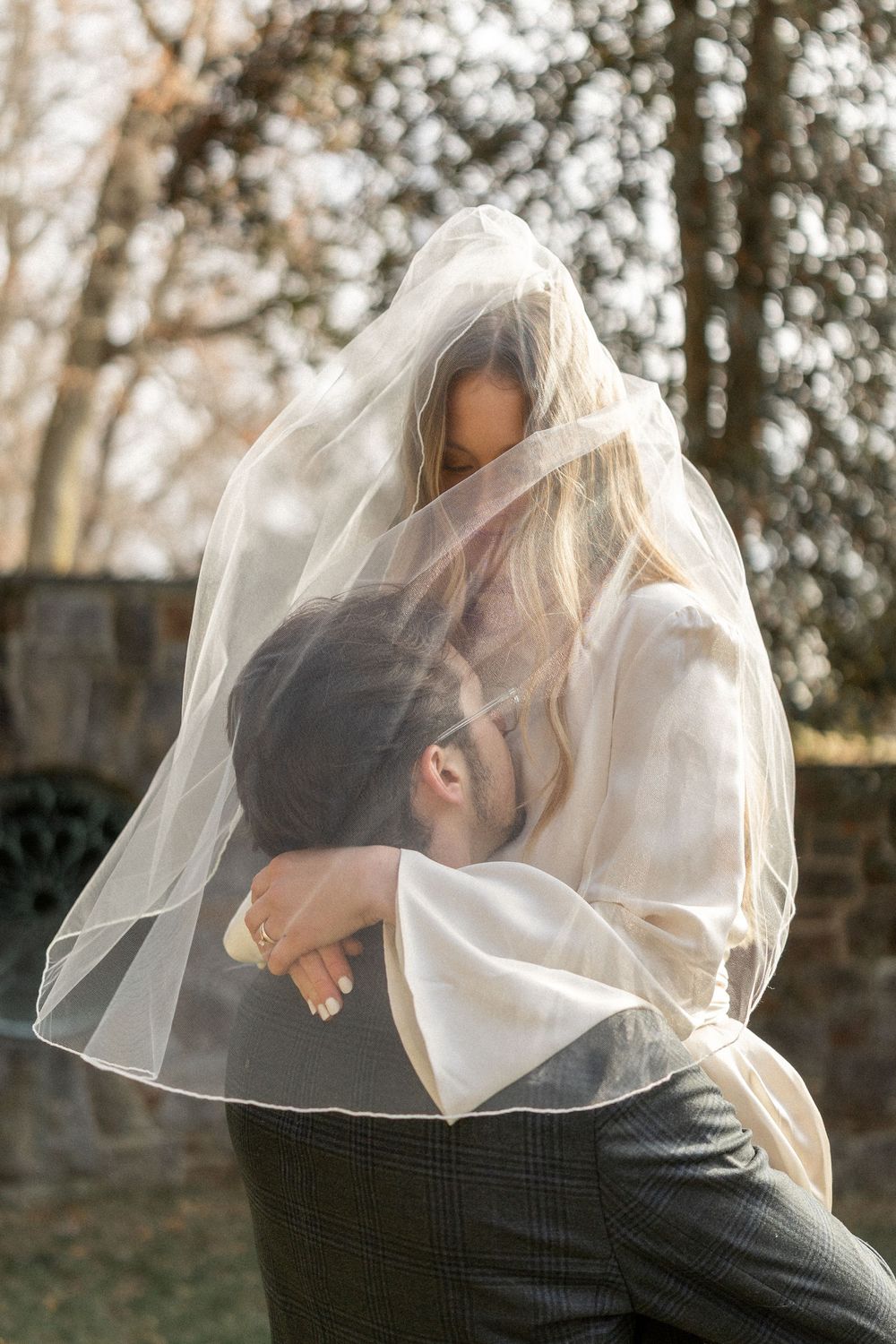A romantic couple embraces under a delicate white veil in a natural outdoor setting during golden hour.