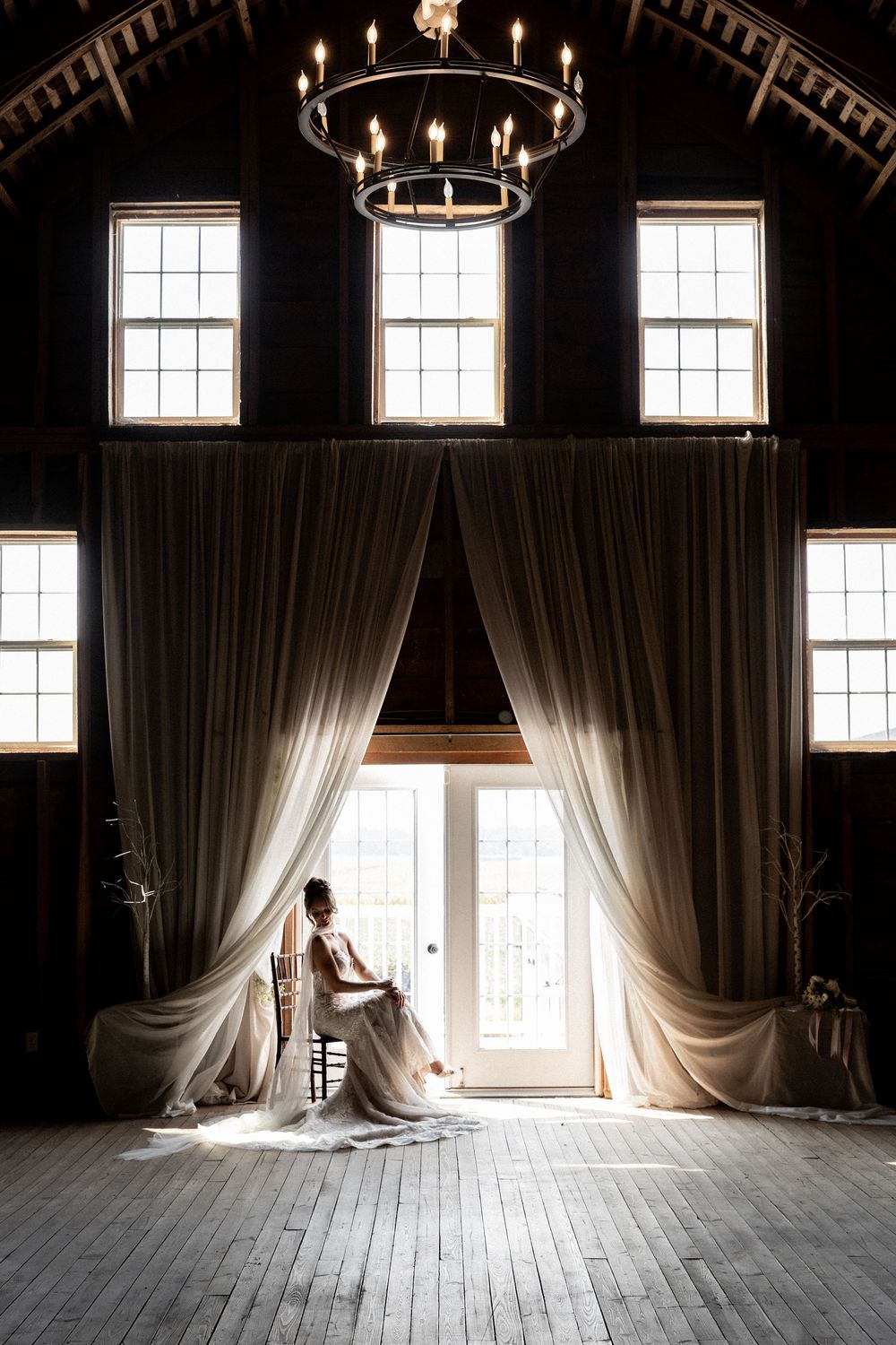 Person sits silhouetted in sunlit wooden room with tall windows and flowing curtains in a moody atmosphere.
