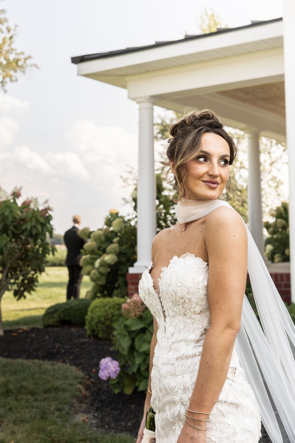 A bride in a detailed lace wedding dress poses on a white columned porch with manicured gardens in the background.