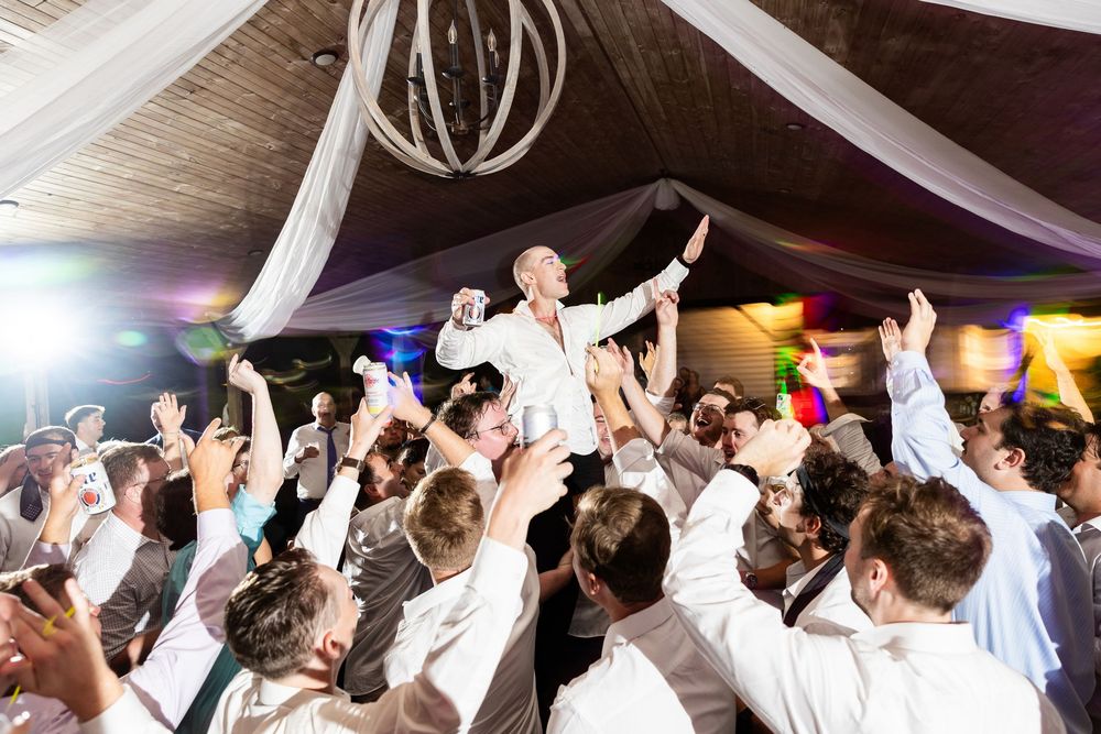 Wedding reception crowd dancing with hands raised on a dance floor under decorative lighting.