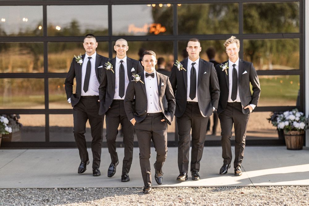 A group of groomsmen in black suits walk together outdoors during a wedding photoshoot.